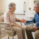 Elderly woman comfortably using a bedside commode chair with caregiver assistance in a well-lit bedroom.